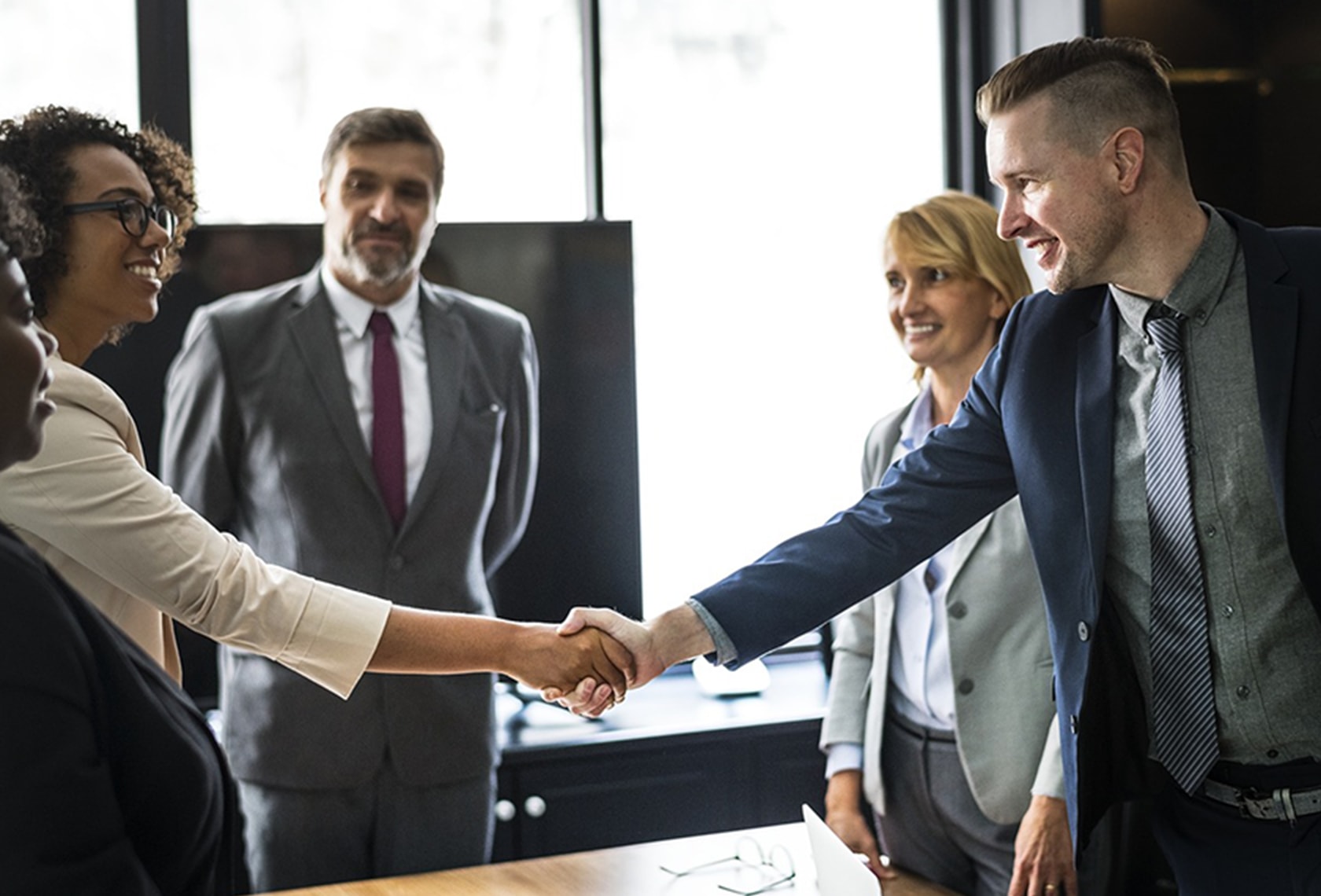 基本のビジネス英語「挨拶」について解説する記事中のイメージ画像です。数人のビジネスパーソンが輪になって立っており、手前の2人が笑顔で握手しています。